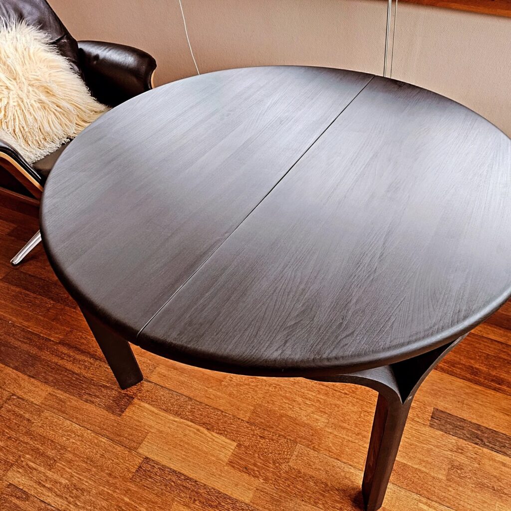 Rundt spisebord før og efter indfarvning – fra lys bøg til mørk elegant finish. Bordet står i en hyggelig stue med stole omkring.